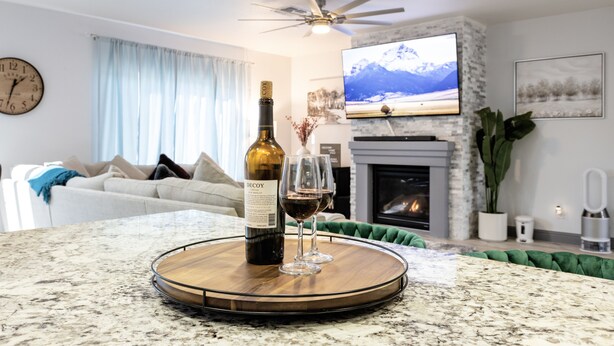A living room with a fireplace, a television mounted above it, and a dining area with a bottle of wine and two glasses on a wooden tray. - Family Pool Home – Saltwater Pool & Fire Pit – 2 King Suites – Sleeps 12 (Elk Grove)