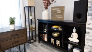 Un soggiorno moderno con una scrivania in legno scuro, una libreria, un vaso bianco con rami secchi, un cartello di benvenuto, una statua di Buddha e un altoparlante nero.