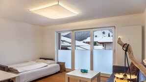 A room with a bed, a table, a sofa, and a lamp. There are large windows with a view of a snowy landscape. - Apartment "Haus Excelsior Top 30" with Mountain View, Balcony & Wi-Fi (Seefeld in Tirol)