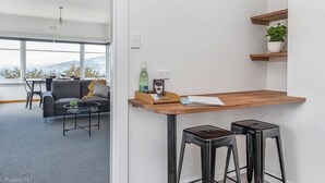 Een moderne woonkamer met een bank, salontafel en een eetgedeelte met stoelen. Er zijn twee barkrukken en een houten tafel met een fles en een notitieboekje.