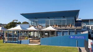 Een modern gebouw met een glazen gevel, een bord met de tekst 'Bellerive Yacht Club' en buiten zitplaatsen.