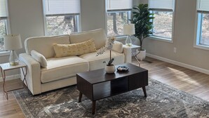A spacious living room with a large window, a white sofa, a dark coffee table, and a modern chandelier.