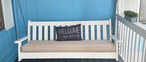 A porch with a white swing bench, a welcome sign, and a logo on the wall.