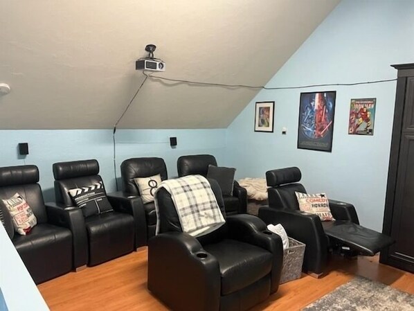 Una sala de cine en casa con sillones reclinables de cuero negro, un proyector instalado en el techo y carteles en la pared.