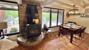 A cozy living room with a stone fireplace, a dining table with chairs, and large windows offering a view of the outdoors. - Private Forest Lodge w/Game House, Spa, mins to DT (Prescott)
