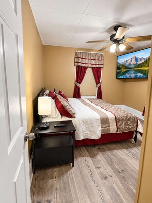 A bedroom with a bed, a nightstand, a remote control, a lamp, a ceiling fan, and a painting on the wall. - High Ceilings | King Beds | 85” TV | Near Downtown (Winston-Salem)
