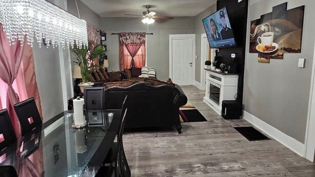 A modern living room with a glass dining table, black sofa, and a TV mounted on the wall. - High Ceilings | King Beds | 85” TV | Near Downtown (Winston-Salem)