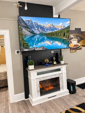 A living room with a fireplace, a mounted television displaying a mountain lake, and a coffee-themed artwork. - High Ceilings | King Beds | 85” TV | Near Downtown (Winston-Salem)