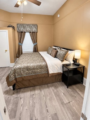 A bedroom with a bed, a nightstand, a ceiling fan, and a window with curtains. - High Ceilings | King Beds | 85” TV | Near Downtown (Winston-Salem)