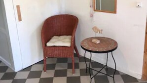 Une pièce avec un sol à carreaux, une chaise en osier avec un coussin, une petite table ronde ornée d’un objet décoratif et un miroir au mur.