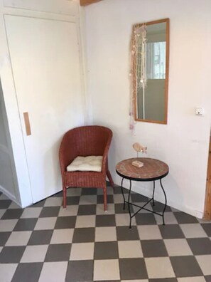 Una habitación con piso a cuadros, una silla de mimbre con cojín, una pequeña mesa redonda con un objeto decorativo y un espejo en la pared.