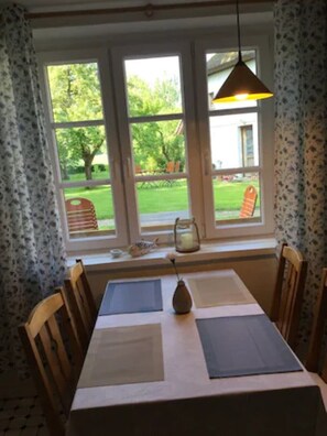 Un comedor con una mesa dispuesta para dos, un florero con flores y vistas a un jardín a través de amplias ventanas.