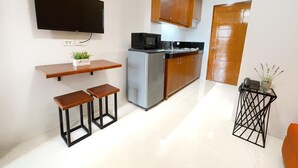 Una cocina compacta con refrigerador, microondas y armarios de madera. Hay un pequeño comedor con taburetes y un estante con una planta.