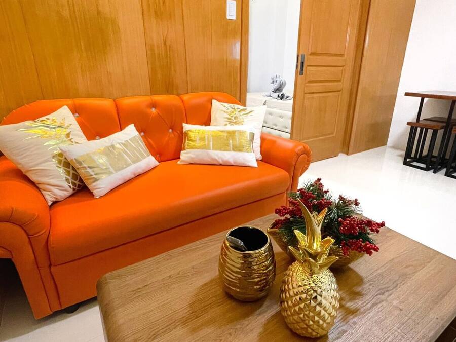 Un divano arancione vivace con cuscini decorativi, un tavolino di legno con un'ananas e un vaso, e una porta di legno sullo sfondo.