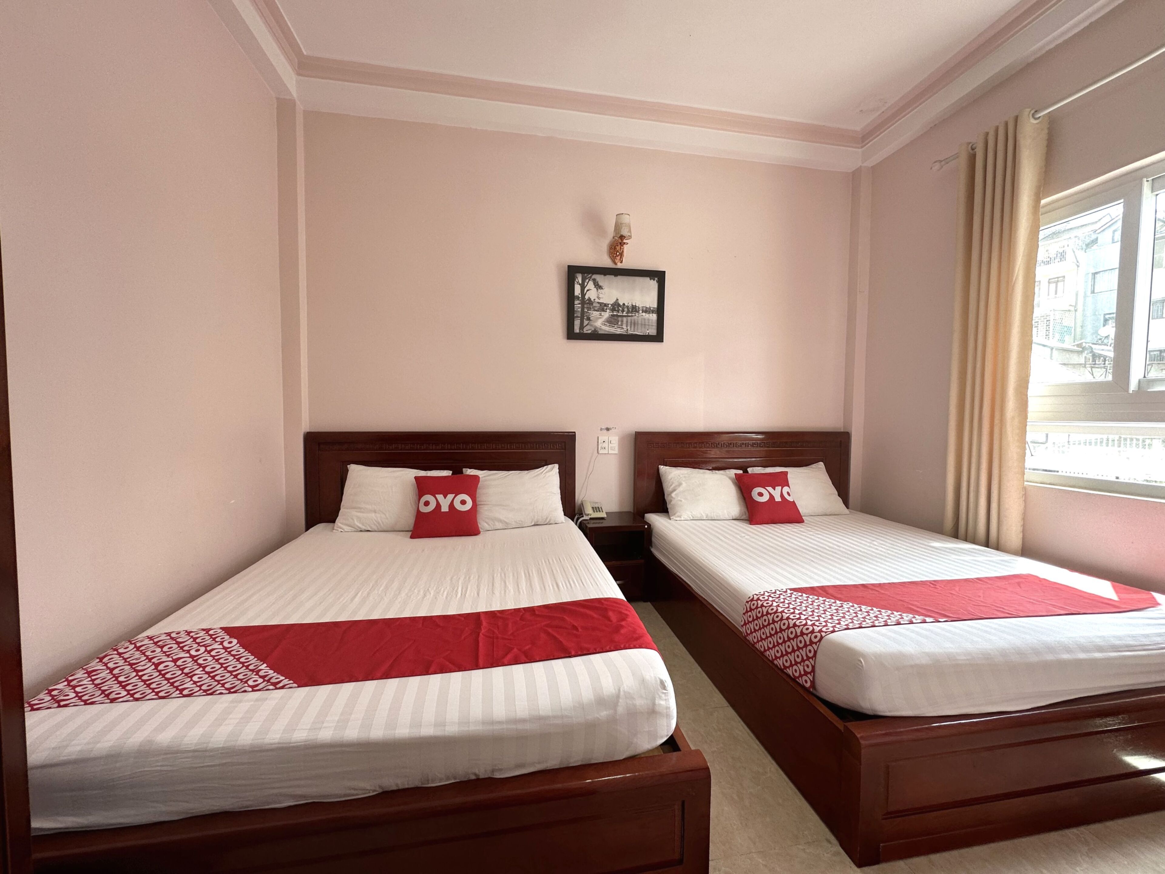 Hotelkamer met twee bedden, elk voorzien van witte beddengoed en rode accenten, een houten hoofdbord, een nachtkastje, een wandlamp en een raam met gordijnen.
