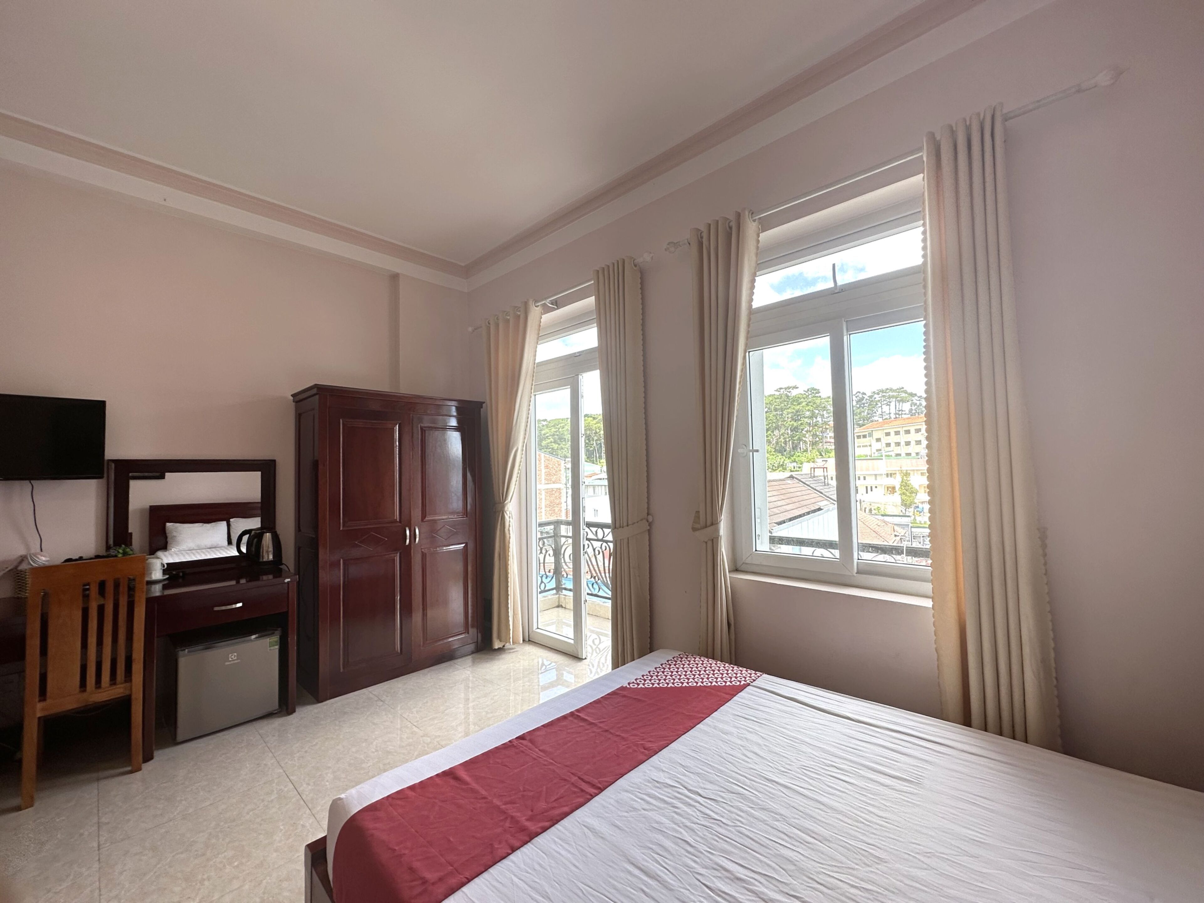 Una camera d'albergo con un letto, una scrivania, un armadio, una televisione e un balcone con vista su edifici e vegetazione.