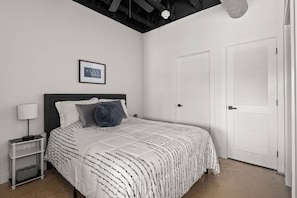 Quarto com cama, mesa de cabeceira, citação motivacional emoldurada e porta branca.