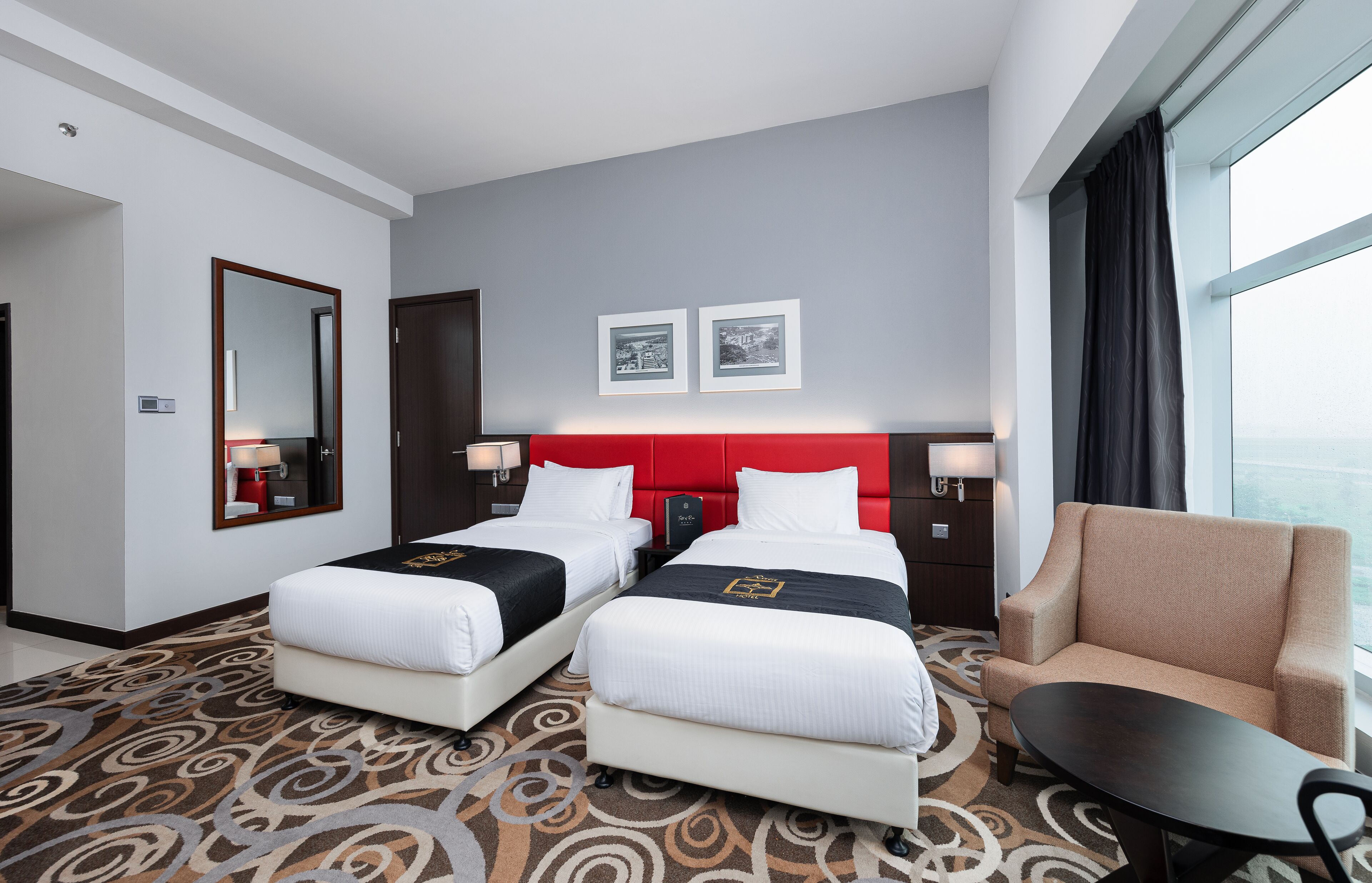 Habitación de hotel con dos camas, cabecera roja, sofá, mesita y un ventanal con vistas.