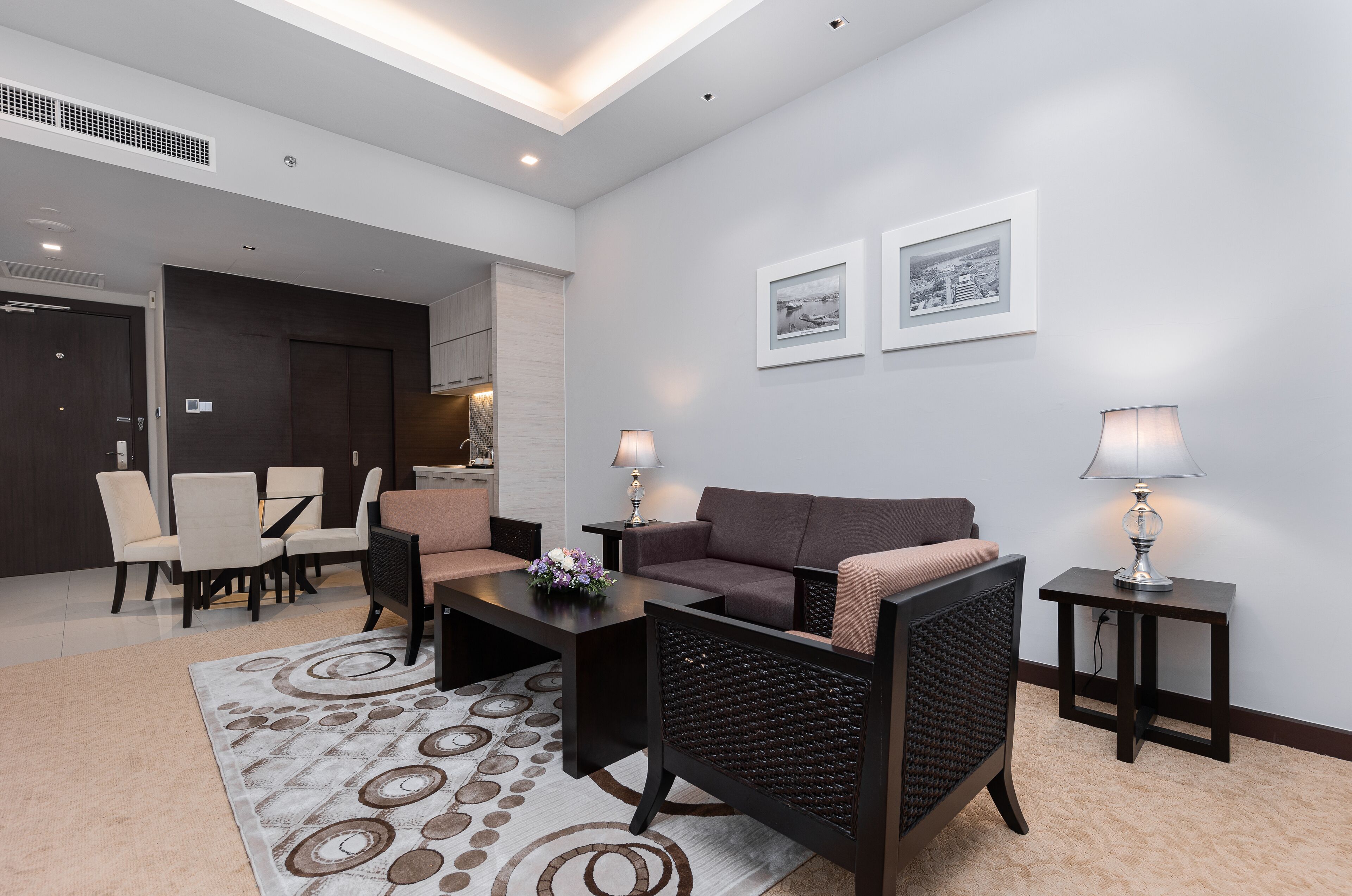 A modern living room with a sofa, armchairs, a coffee table, and a rug. There are two side tables with lamps, a dining area with chairs, and a kitchen area in the background.