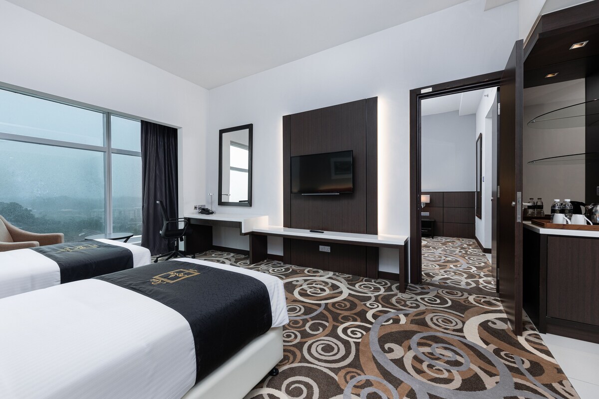 A hotel room with two beds, a TV, a desk, and a view of the city.