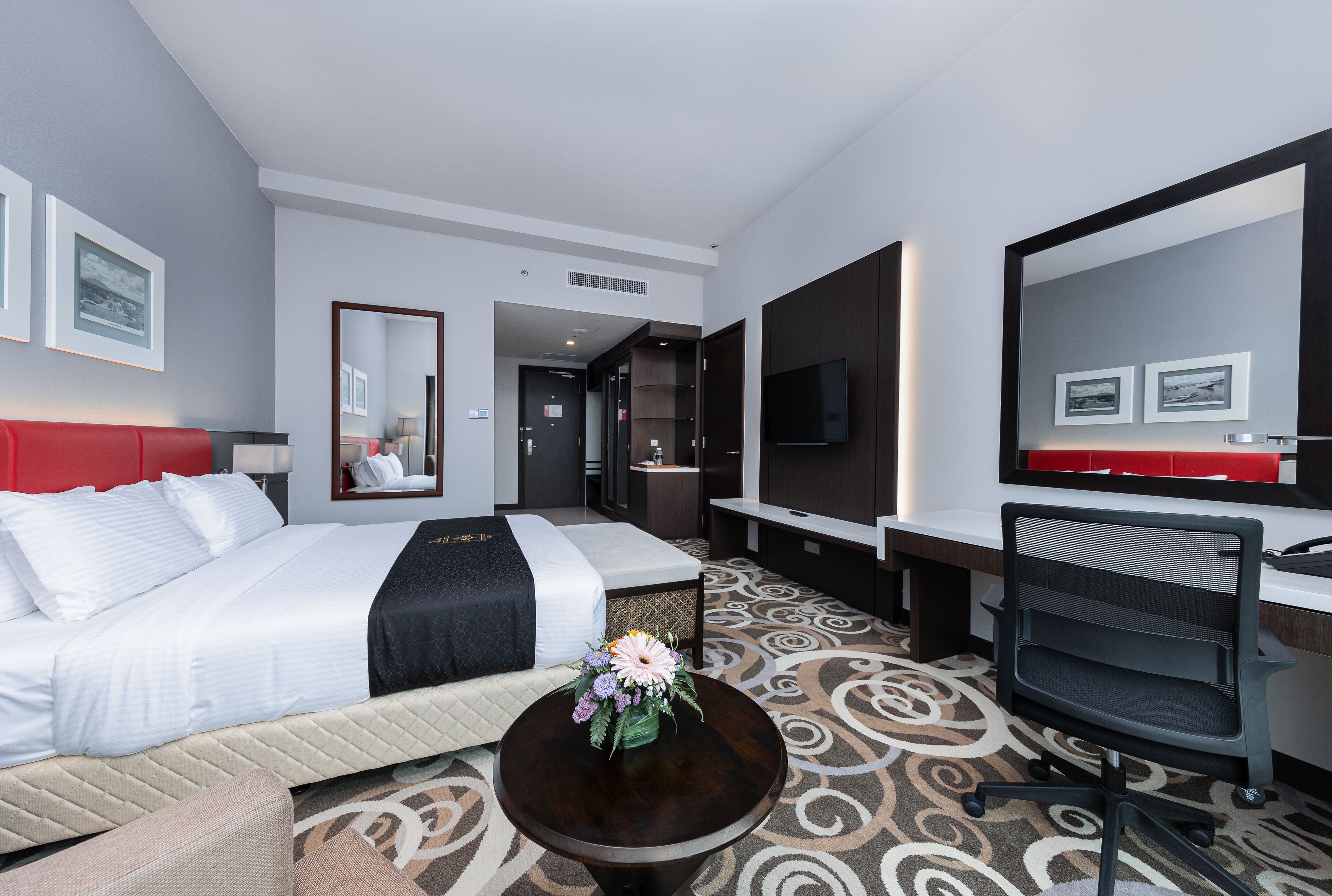 Quarto de hotel com cama, escrivaninha, cadeira, televisão e espelho.