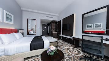 Quarto de hotel com cama, escrivaninha, cadeira, televisão e espelho.