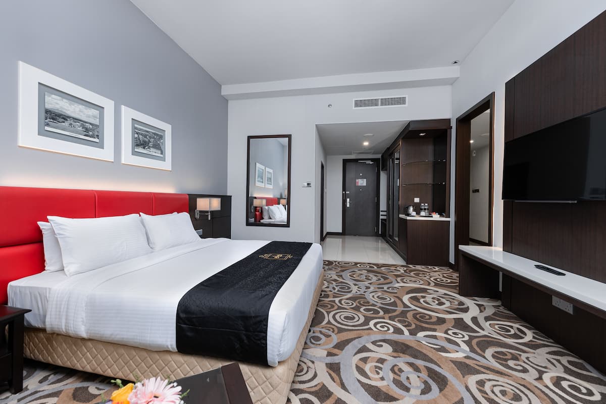 A hotel room with a red headboard, a large bed, a TV, a mirror, and a bathroom visible through an open door.