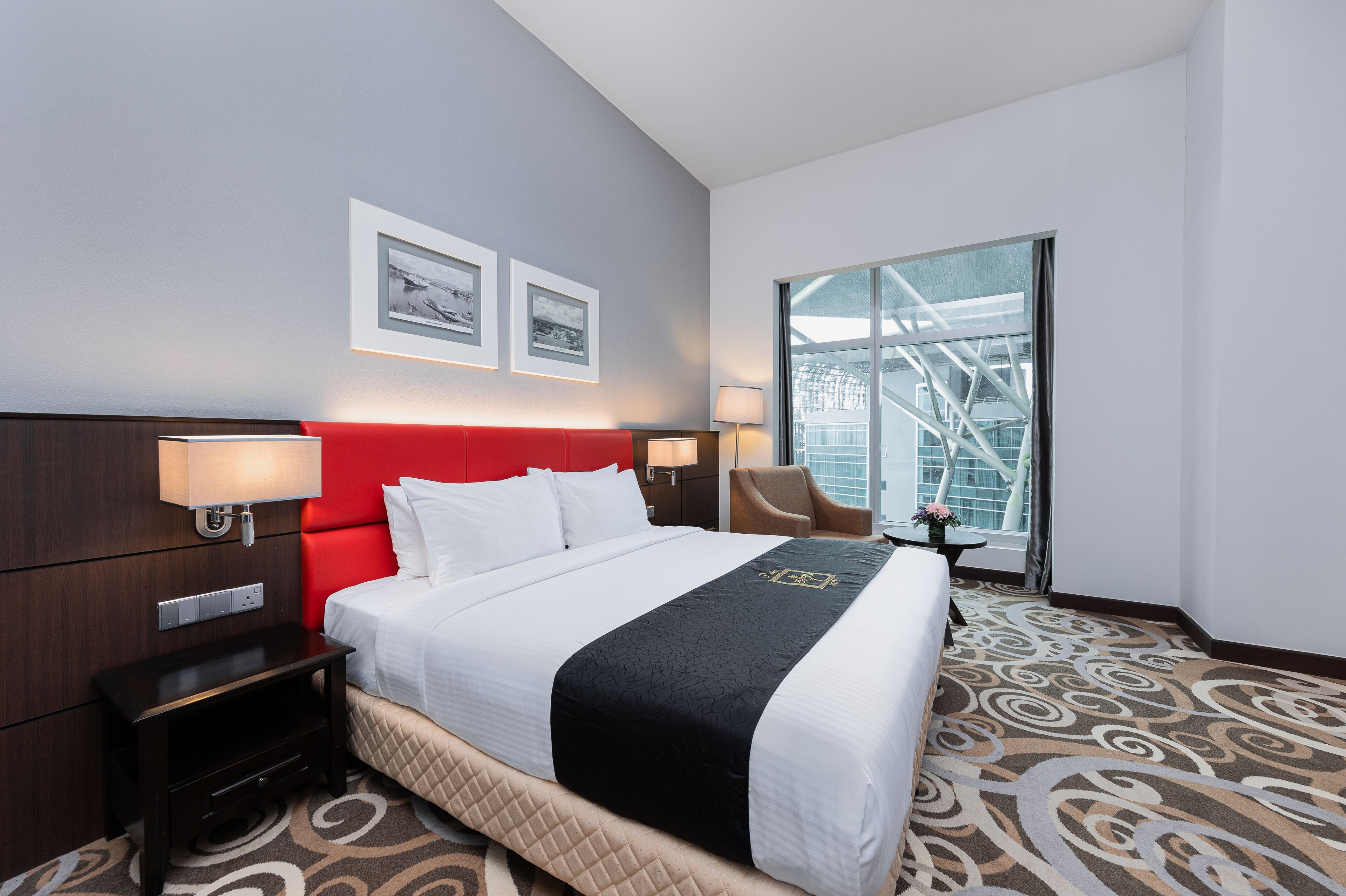 A hotel room with a large bed, a red headboard, two framed pictures on the wall, a window with a view of a building, and a carpet with a swirl pattern.