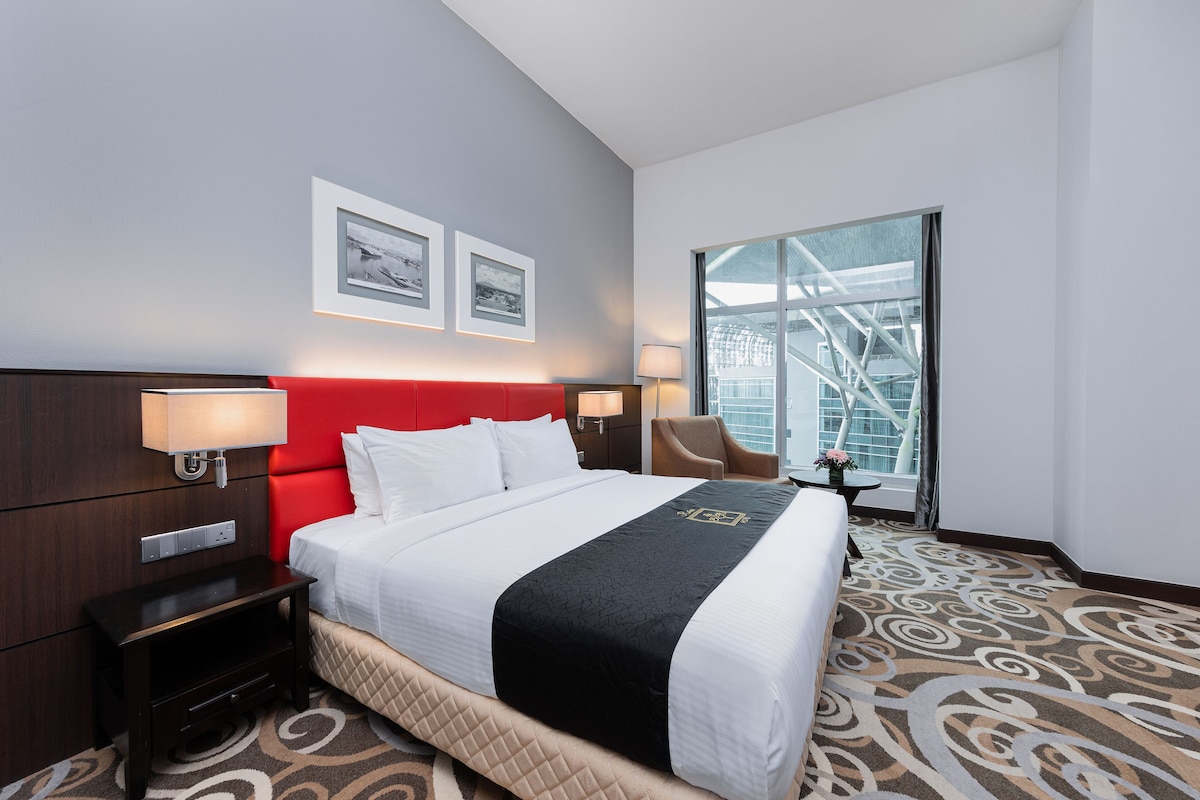 A hotel room with a large bed, a red headboard, two framed pictures on the wall, a window with a view of a building, and a carpet with a swirl pattern.