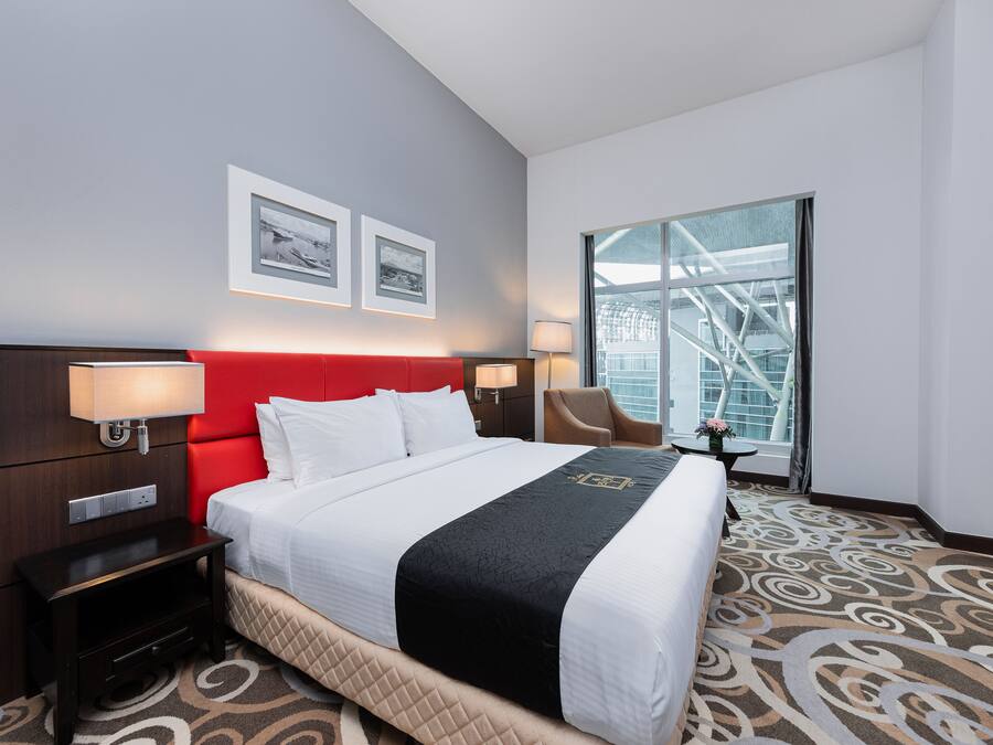 A hotel room with a large bed, a red headboard, two framed pictures on the wall, a window with a view of a building, and a carpet with a swirl pattern.
