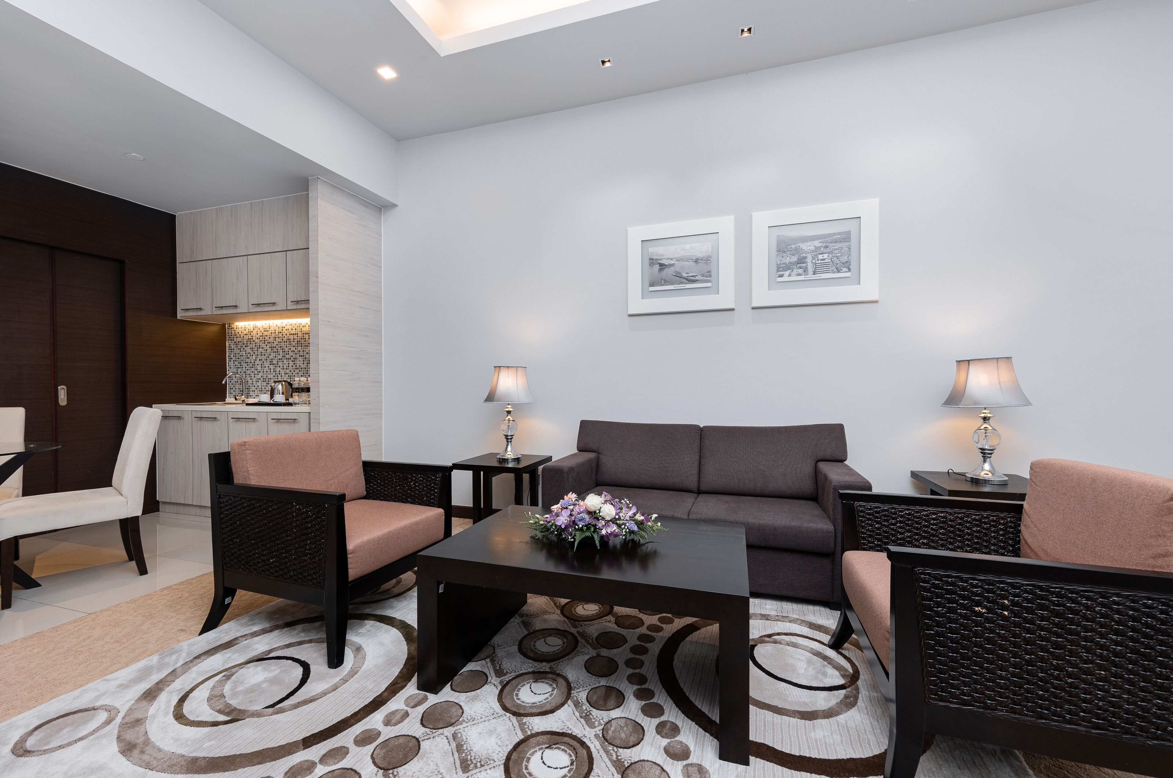 Una sala de estar moderna con un sofá, dos sillones, una mesa de centro y dos mesas auxiliares con lámparas. Se ve una cocina al fondo, con armarios y una encimera. Dos cuadros enmarcados decoran la pared.