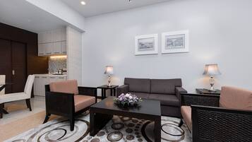 Una sala de estar moderna con un sofá, dos sillones, una mesa de centro y dos mesas auxiliares con lámparas. Se ve una cocina al fondo, con armarios y una encimera. Dos cuadros enmarcados decoran la pared.