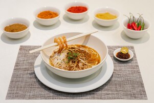 Un tazón de sopa de fideos con palillos, rodeado de pequeños recipientes con condimentos.