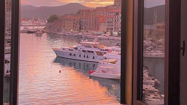A view from a window overlooking a marina with boats and buildings during sunset. - The Rooms by the Sea (Portoferraio)