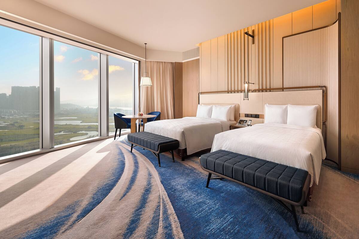 A hotel room with two beds, a large window with a city view, a small table, and a bench.
