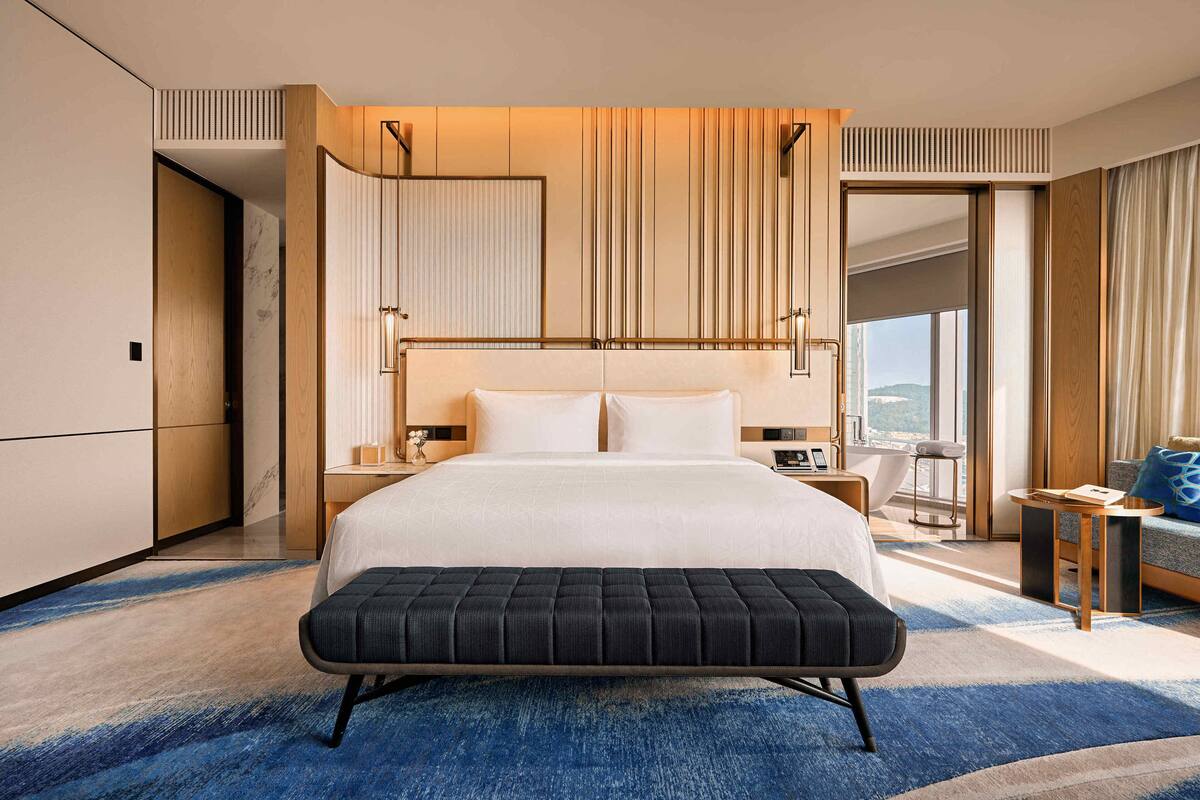 A modern hotel room with a large bed, a bench, a nightstand, a sofa, and a view of the city.