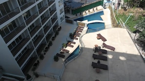 Área de piscina no terraço com espreguiçadeiras e piscina, rodeada por um edifício com vários andares e varandas.