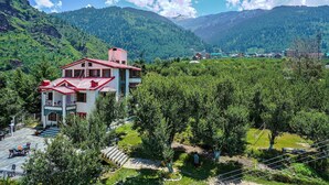 Un bâtiment à deux étages, avec un toit rouge, entouré d’une végétation luxuriante et avec des montagnes en arrière-plan.