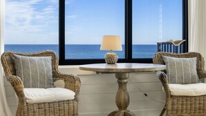 Une pièce avec deux fauteuils en osier, une table ronde avec une lampe, et une vue sur la mer grâce à de grandes fenêtres.