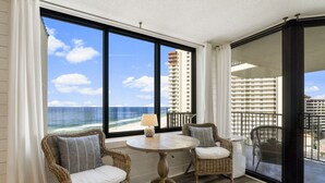 Un balcon avec des fauteuils en osier et une table, d’où l’on peut admirer une plage et des immeubles de grande hauteur.