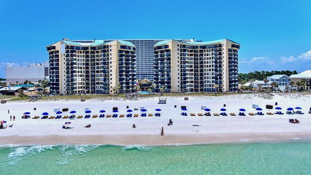 Un hôtel en bord de mer composé de plusieurs immeubles de grande hauteur, une plage de sable fin avec des transats et des parasols, et une eau turquoise et cristalline.