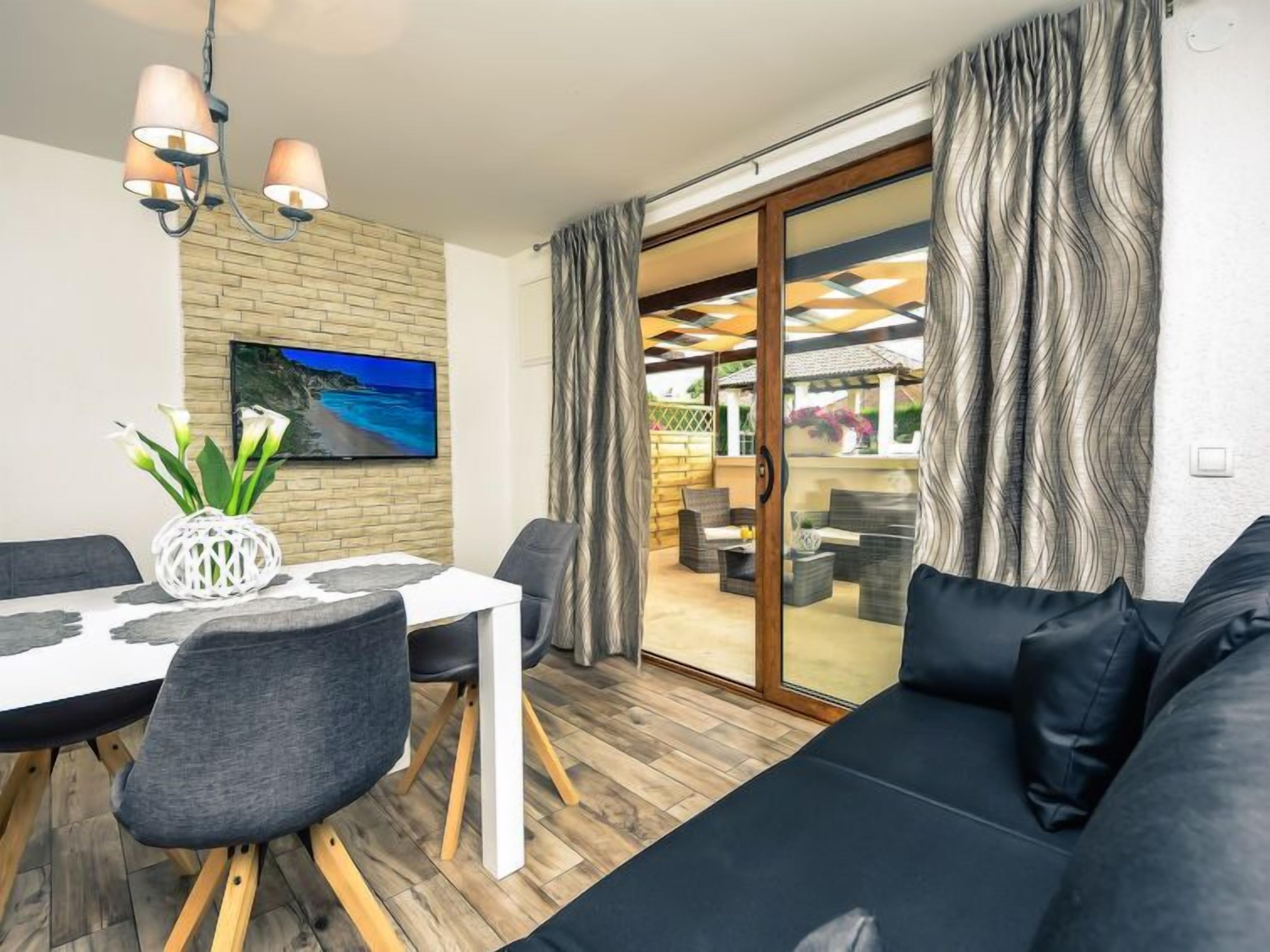 Una sala moderna con comedor, un sofá, un televisor montado en una pared de ladrillo y vistas a través de una puerta de cristal.