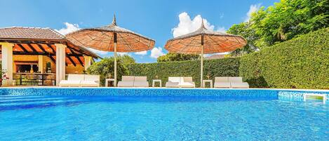A swimming pool with lounge chairs and umbrellas, surrounded by a hedge and a building with a tiled roof.