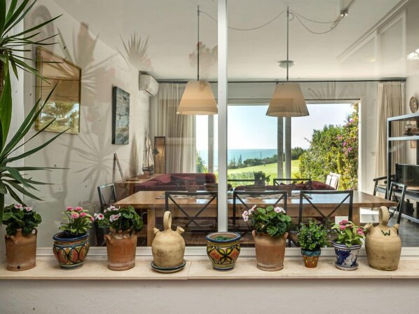 A spacious dining area with a large table, multiple chairs, and a view of the ocean. There are several potted plants on the windowsill.