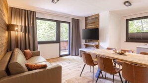 A modern living room with a sofa, a dining table with chairs, a flat-screen TV mounted on a wooden wall, and large windows with curtains. - Suite mit 2 SZ & Wellnessbereich by Interhome (Saalbach-Hinterglemm)