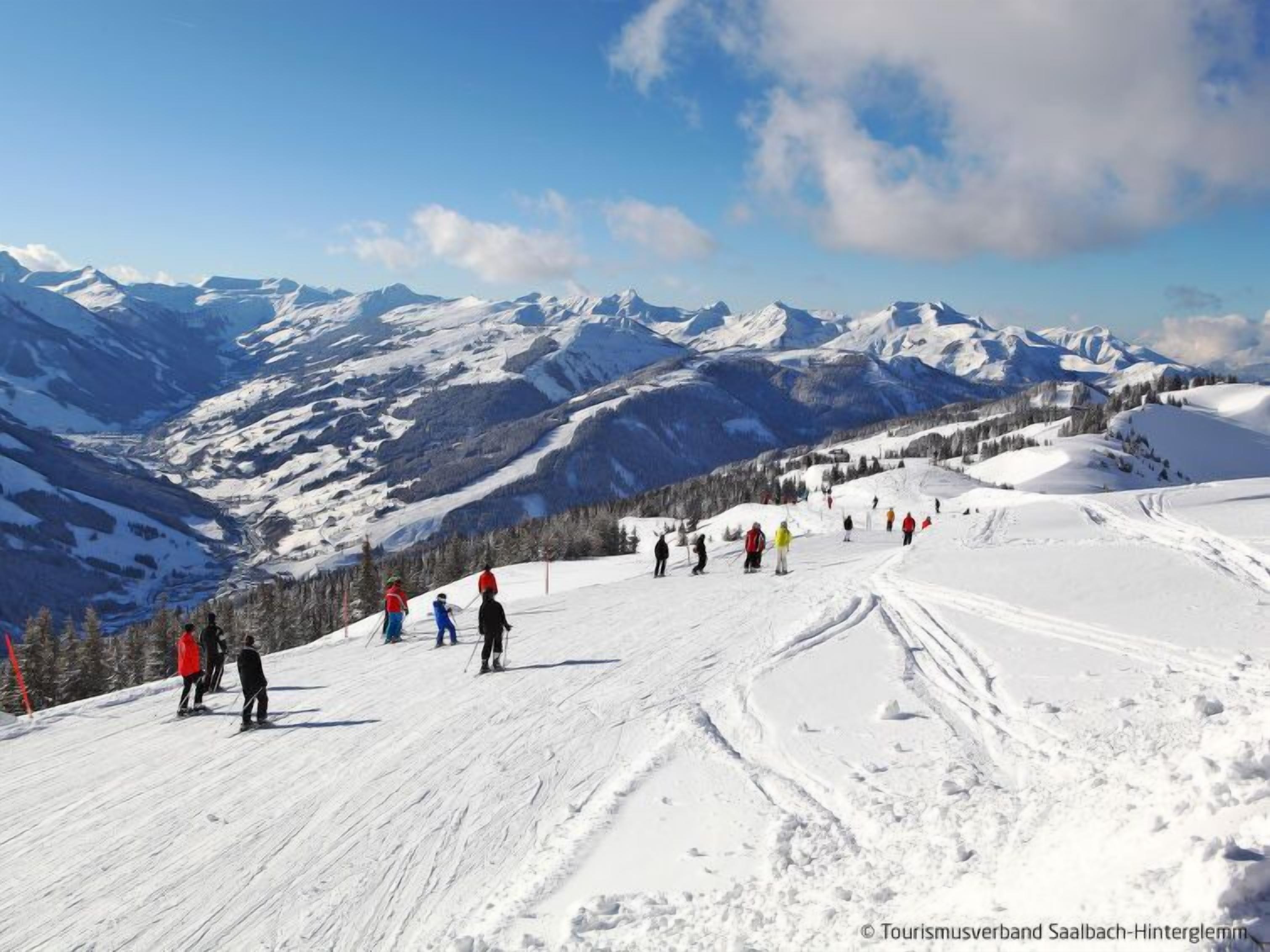 A snowy ski slope with skiers and snowboarders, surrounded by mountains and clear skies.