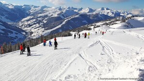 A snowy ski slope with skiers and snowboarders, surrounded by mountains and clear skies.