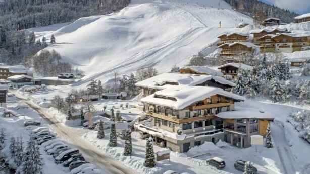 A snow-covered mountain resort with chalet-style buildings, a ski slope, and a parking area.