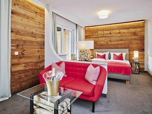 A modern hotel room with a wooden wall, a red sofa, a bed with red bedding, and a glass coffee table with a bottle and glasses.
