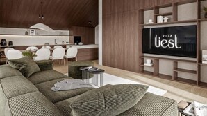 Un salon moderne avec un grand canapé d’angle, un téléviseur à écran plat et une cuisine en arrière-plan.